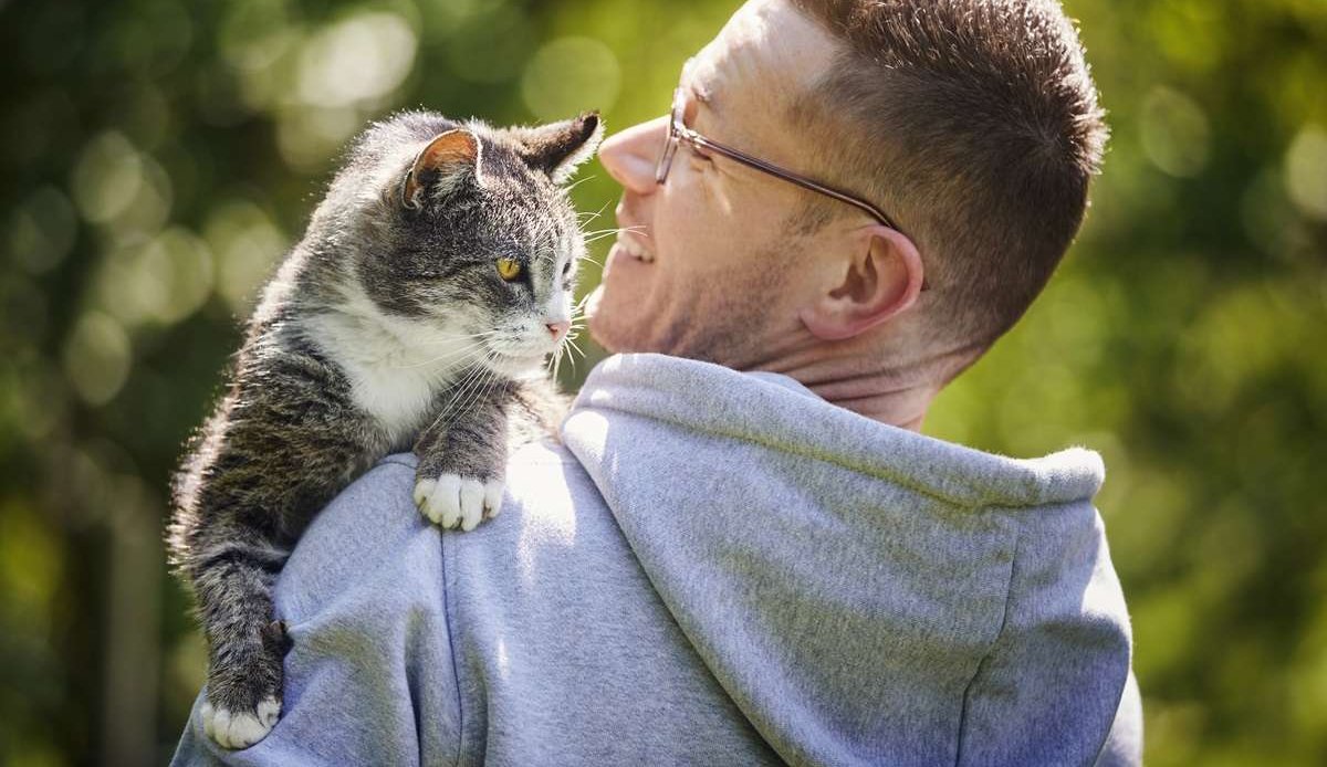 Uomo sorridente che porta in braccio il suo gatto soriano anziano in giardino